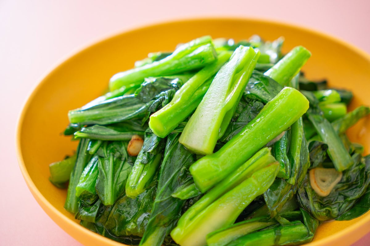 A yellow plate filled with stir-fried leafy green vegetables and slices of garlic against a light pink background.