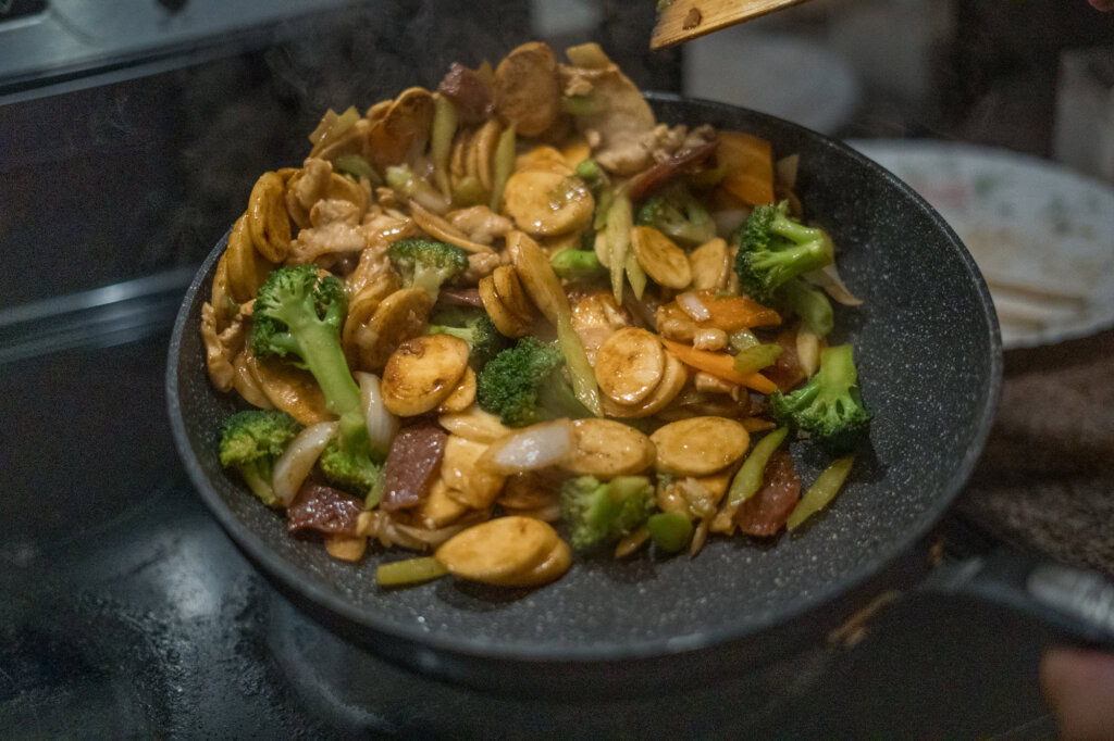 A pan filled with stir-fried vegetables, sliced sausage, tofu, and sauce being cooked on a stovetop.