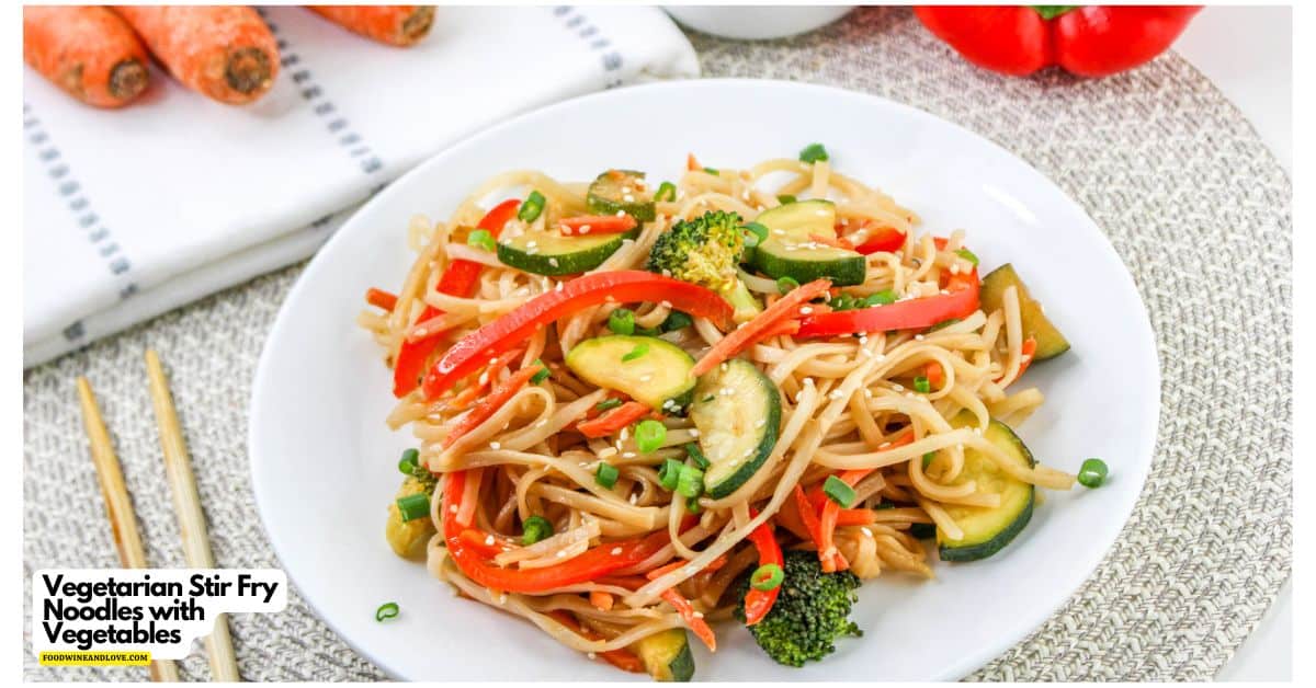 A white plate of vegetarian stir fry noodles with zucchini, broccoli, red bell peppers, and carrots, garnished with green onions. Chopsticks and fresh vegetables are nearby.