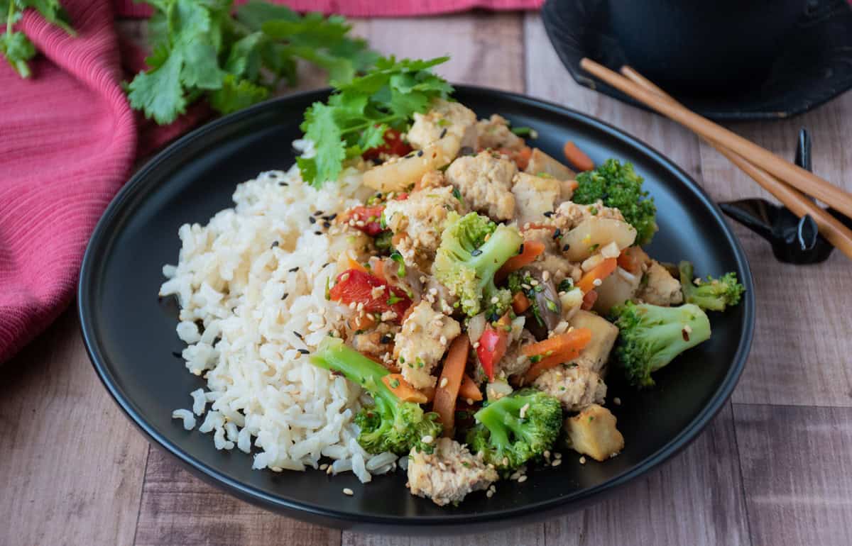 A black plate with white rice and vegetable stir-fry with chicken, garnished with fresh cilantro, on a wooden surface with chopsticks and a red cloth nearby.