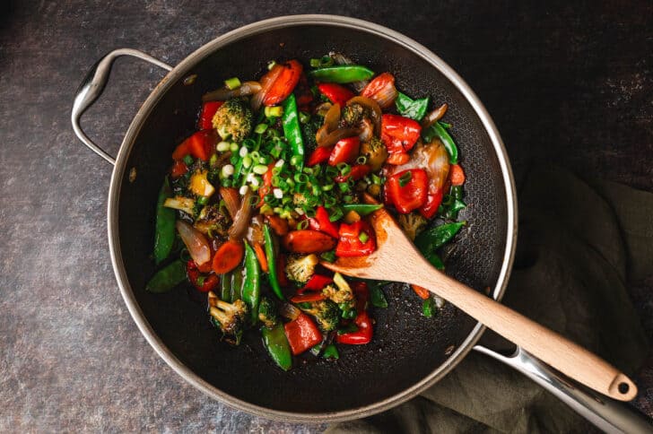 A stir-fry with assorted vegetables, including bell peppers, broccoli, snow peas, and carrots, is being cooked in a large skillet with a wooden spoon.