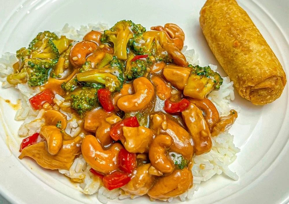 A plate of white rice topped with cashew chicken, broccoli, and red peppers in sauce, served with a fried egg roll on the side.