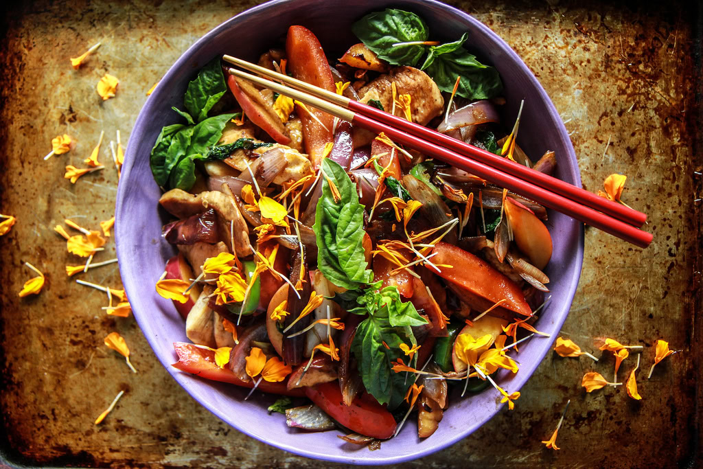 A bowl of stir-fried vegetables and chicken garnished with fresh basil leaves and yellow flower petals, topped with a pair of red and yellow chopsticks.