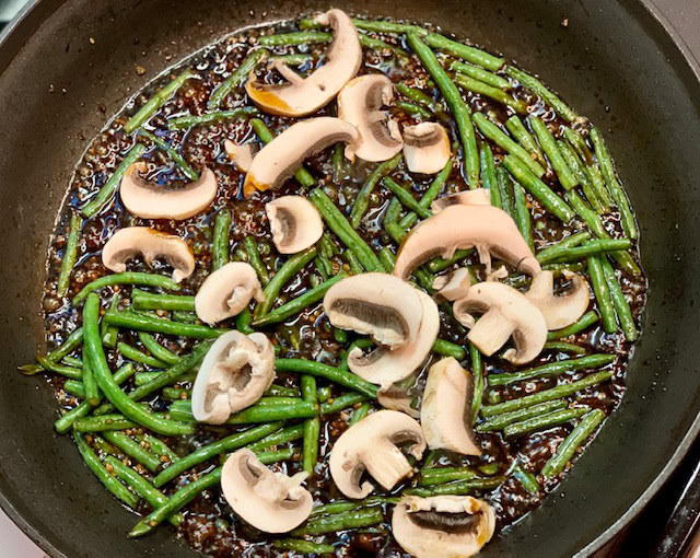 Sliced mushrooms and green beans are cooking in a pan with a dark sauce.