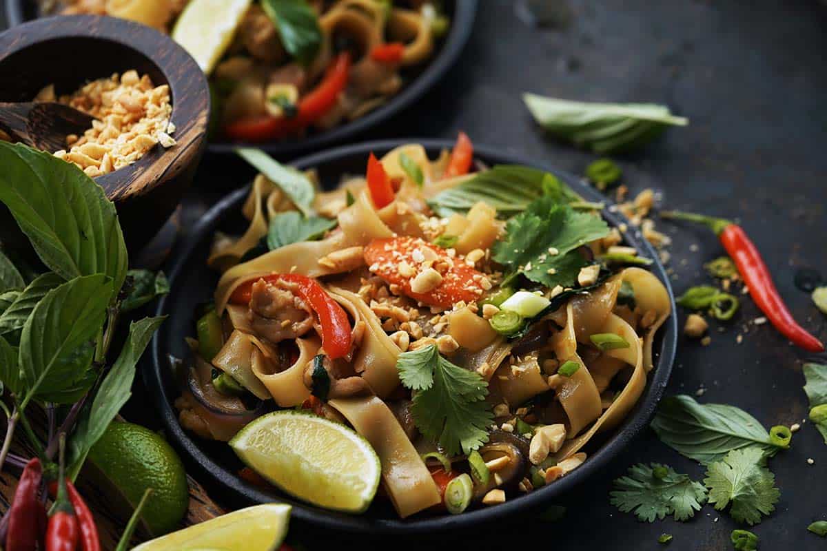 A bowl of stir-fried noodles with red peppers, lime wedges, peanuts, cilantro, and green onions, garnished with sesame seeds; herbs and chilies are scattered around.