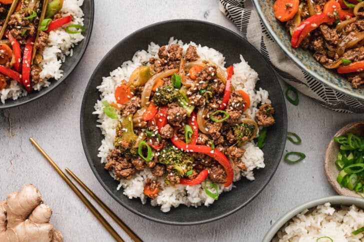 Bowl of white rice topped with stir-fried ground beef, broccoli, red bell peppers, and carrots, garnished with sesame seeds and sliced green onions. Chopsticks and ginger root nearby.