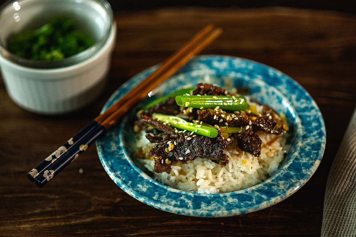 A blue bowl of white rice topped with beef, green vegetables, and sesame seeds, with chopsticks resting on the bowl; a small bowl of herbs is in the background.
