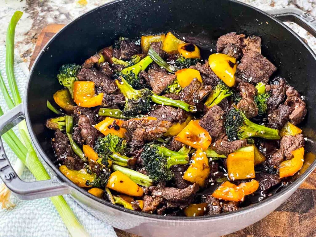 A cast iron pot filled with beef stir-fry, featuring broccoli florets and yellow bell peppers, topped with sesame seeds.