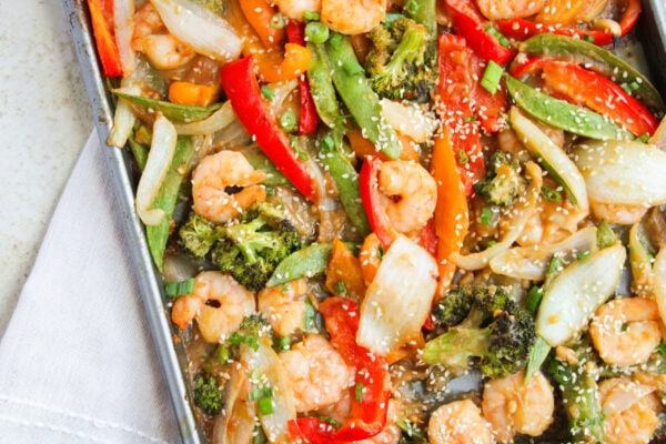 Sheet pan with cooked shrimp, broccoli, snap peas, red bell peppers, onions, and carrots, garnished with sesame seeds and green onions.