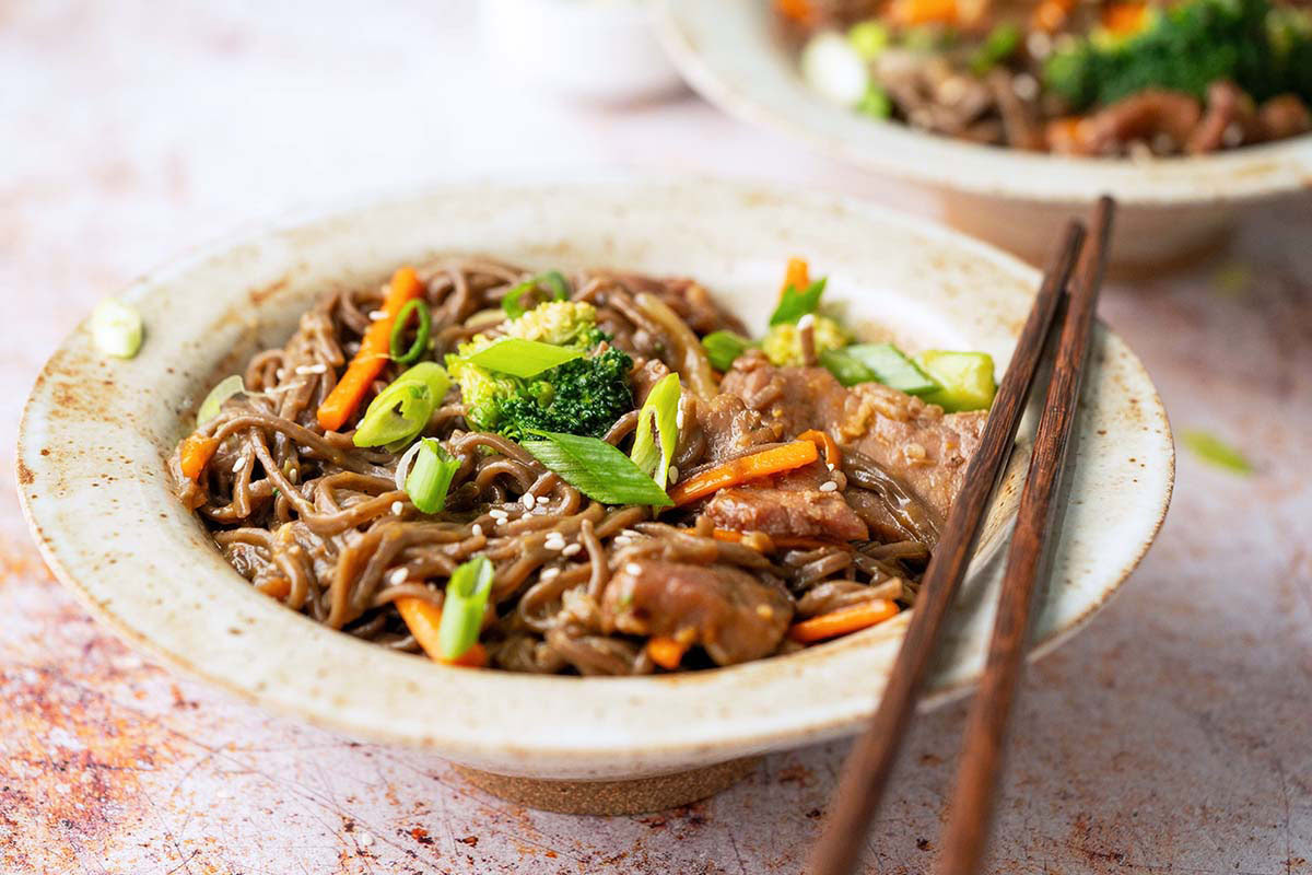 A bowl of soba noodles with sliced beef, broccoli, carrots, and green onions, garnished with sesame seeds and served with chopsticks.