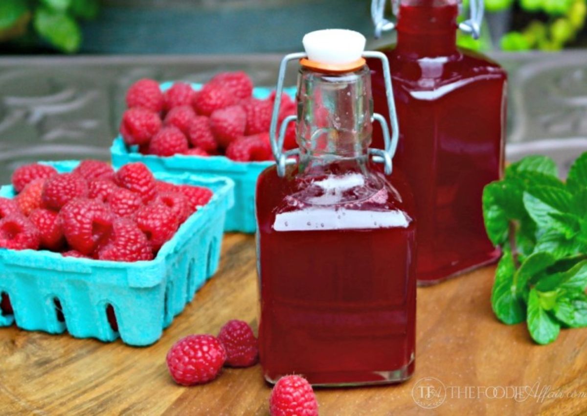 Two glass bottles of raspberry syrup sit on a wooden surface next to cartons of fresh raspberries and mint leaves.