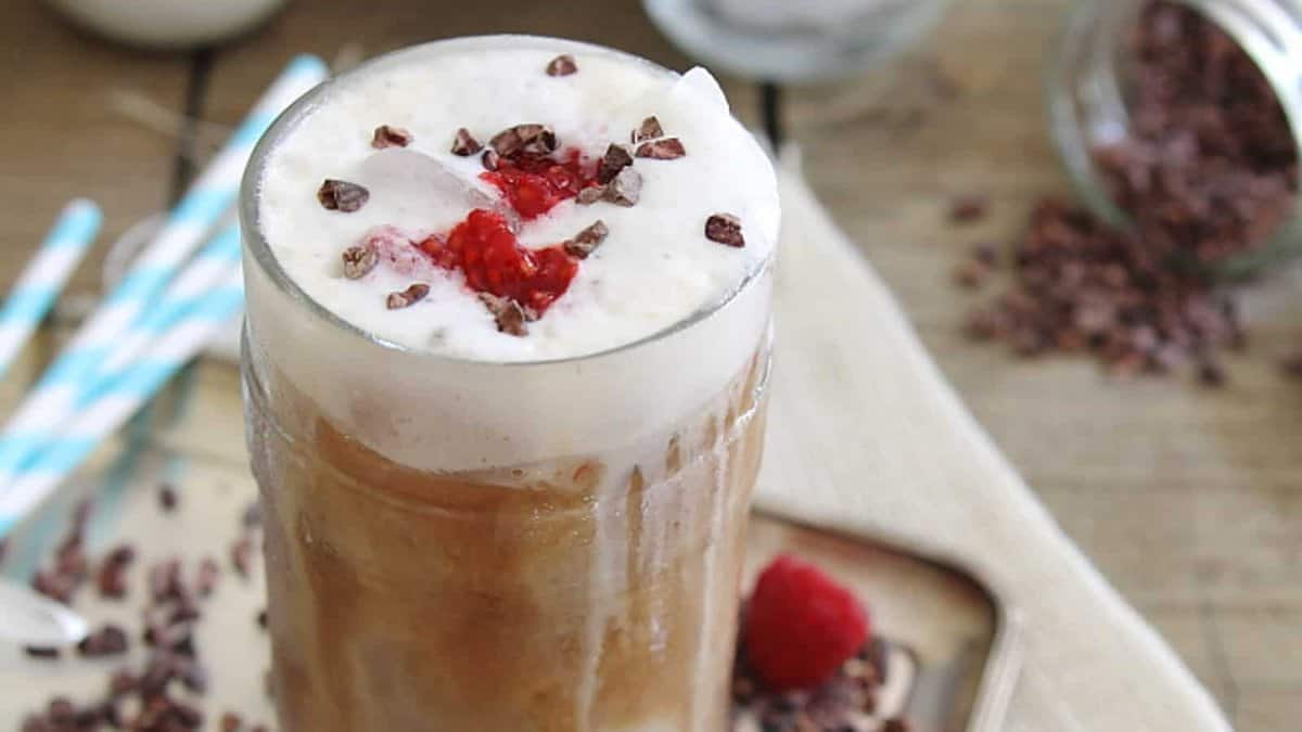 A glass of iced coffee topped with whipped cream, cacao nibs, and a raspberry, with straws and cacao nibs scattered on the table.
