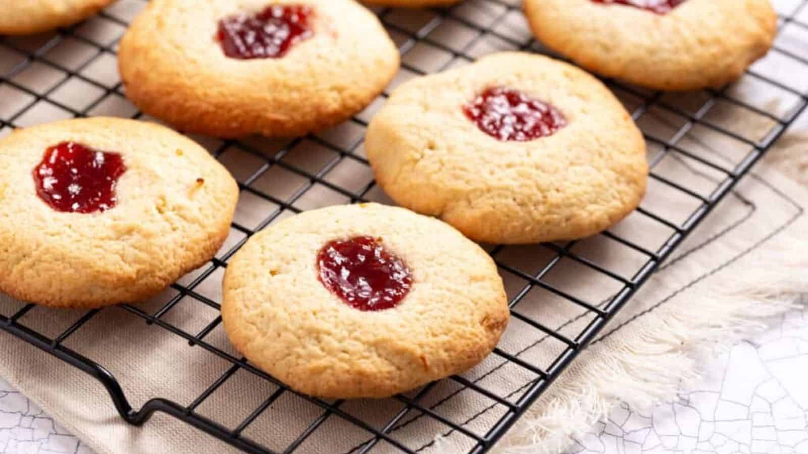Round cookies with a dollop of red jam in the center are cooling on a black wire rack over a beige cloth.