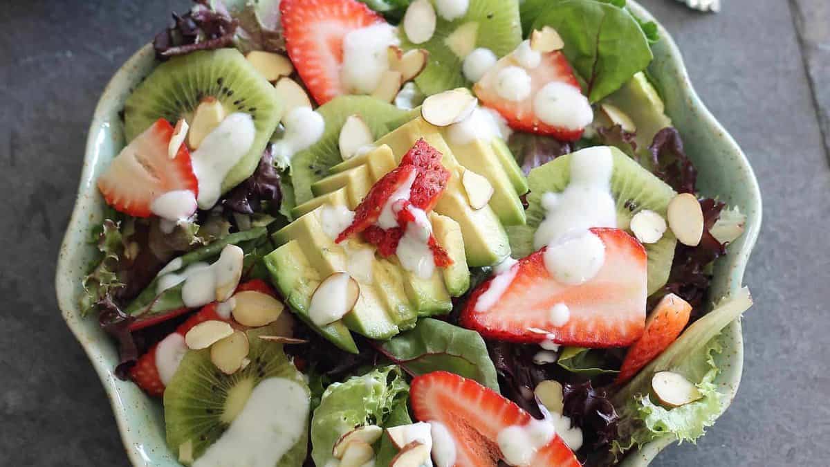 A bowl of mixed greens topped with sliced strawberries, kiwi, avocado, almond slices, and drizzled with a creamy dressing.
