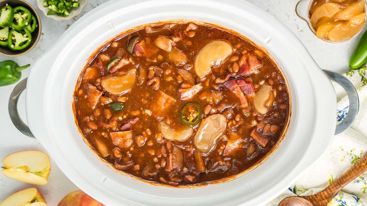 A slow cooker filled with baked beans, large white beans, bacon pieces, and chopped vegetables, surrounded by sliced apples, jalapeรฑos, and a wooden spoon.