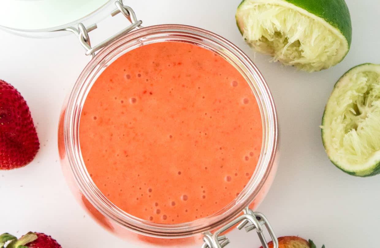A glass jar filled with a smooth, pink strawberry sauce sits next to a halved, juiced lime and fresh strawberries on a white surface.