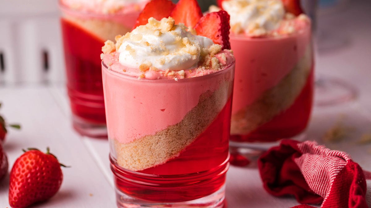 Three glasses of layered strawberry dessert with red jelly, crumb crust, pink mousse, whipped cream, and sliced strawberries, set on a white surface with fresh strawberries nearby.