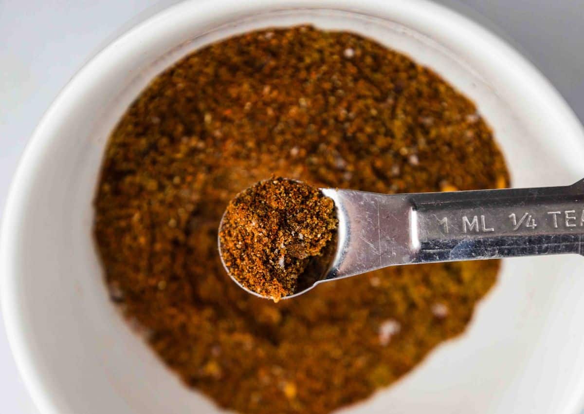 A metal measuring spoon holding a small amount of dark brown spice mix is held over a white bowl filled with the same spice mixture.