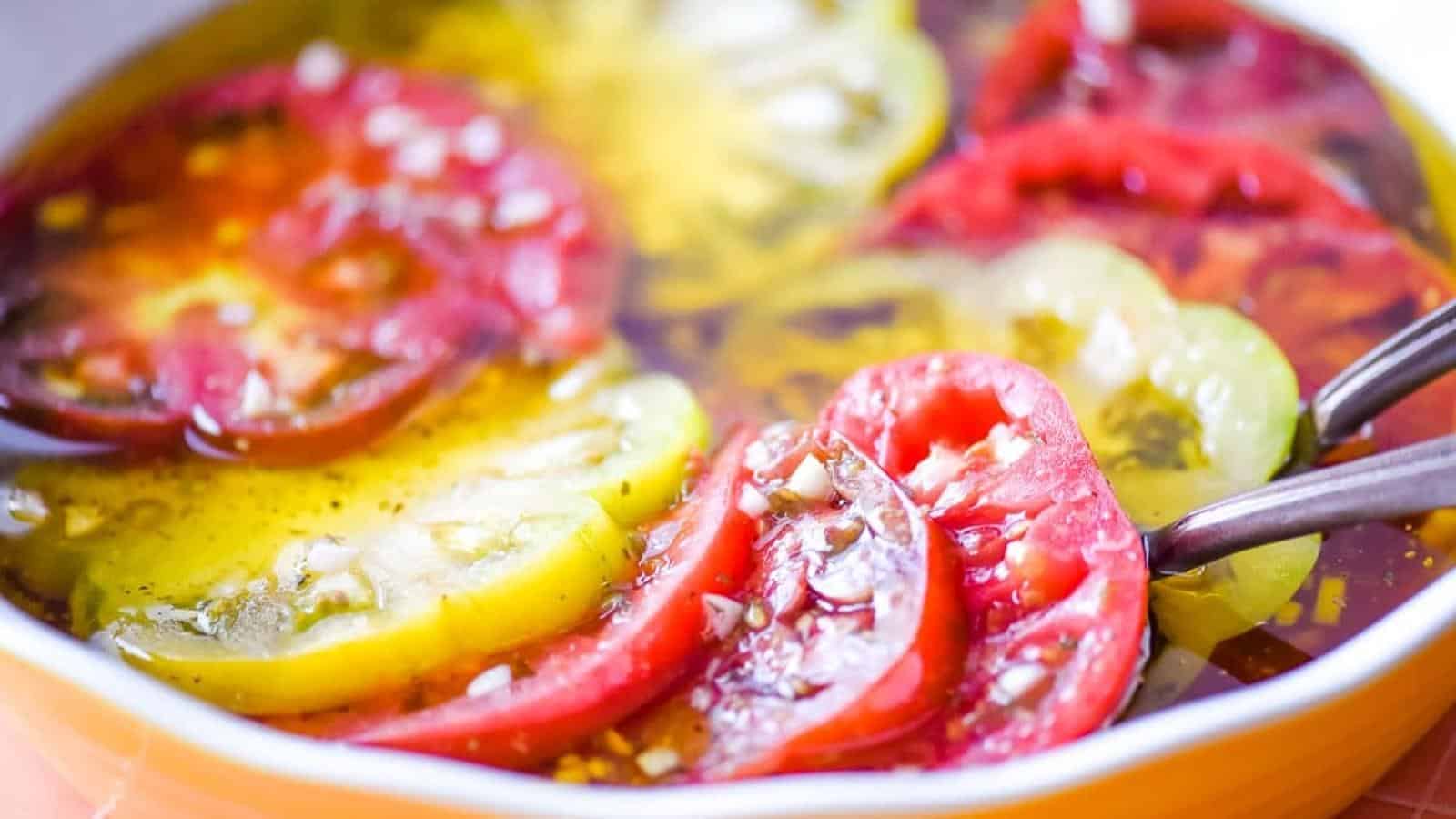 Sliced red and green tomatoes in a shallow dish, topped with minced garlic, herbs, oil, and vinegar, with two metal forks.