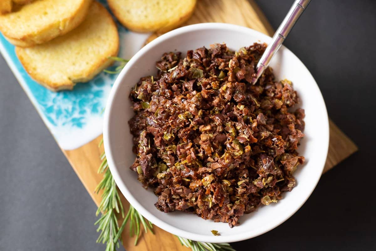 A white bowl filled with chopped olive tapenade sits on a cutting board, with toasted bread slices and a sprig of rosemary nearby.
