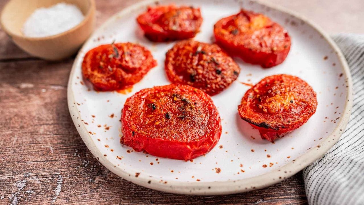 A plate with six slices of roasted tomatoes, lightly browned, next to a small bowl of coarse salt and a striped cloth napkin on a wooden surface.