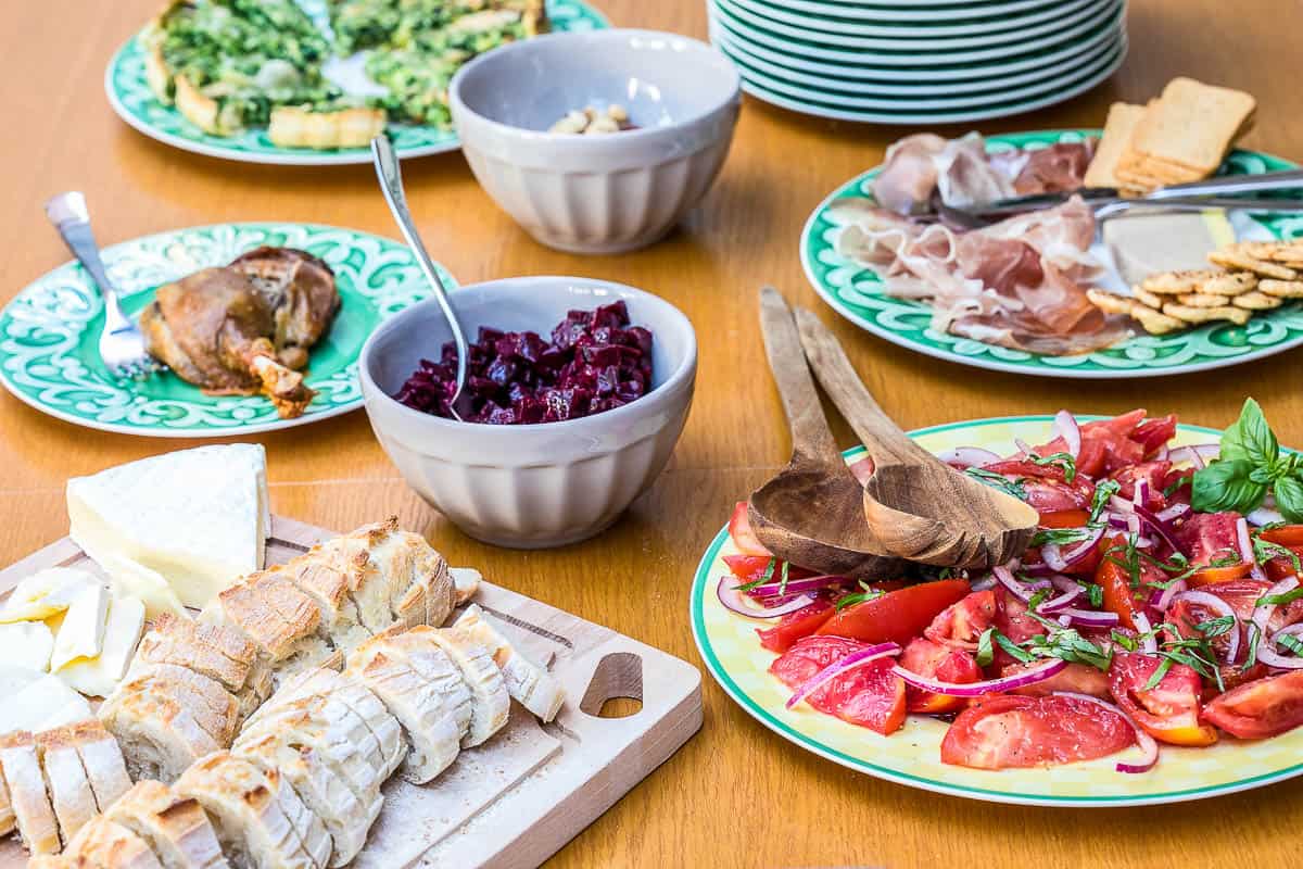 A table set with plates of sliced bread and cheese, tomato salad, cured meats with crackers, cooked chicken, pasta, and a bowl of diced beets.