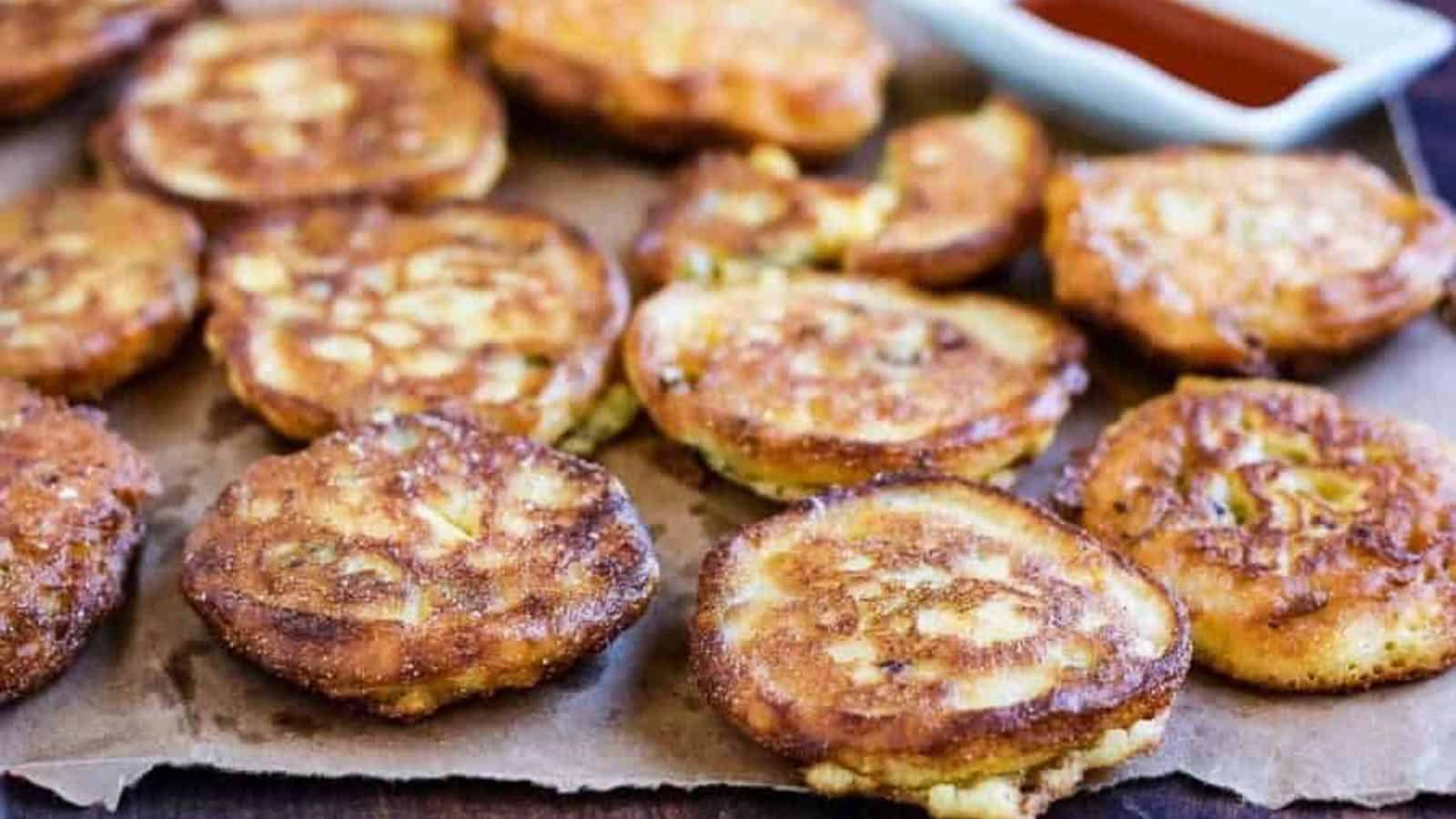 A batch of golden-brown fritters arranged on parchment paper, with a small dish of red dipping sauce in the background.
