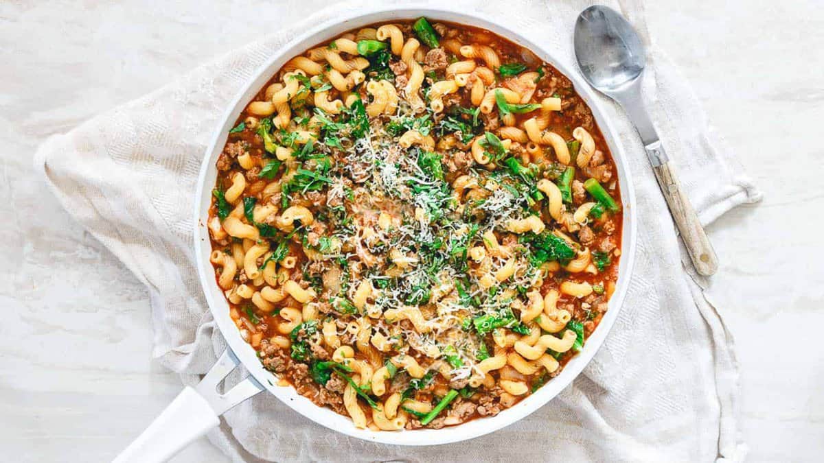 A pan filled with cooked pasta, ground meat, spinach, tomato sauce, and grated cheese, placed on a cloth napkin with a spoon beside it.