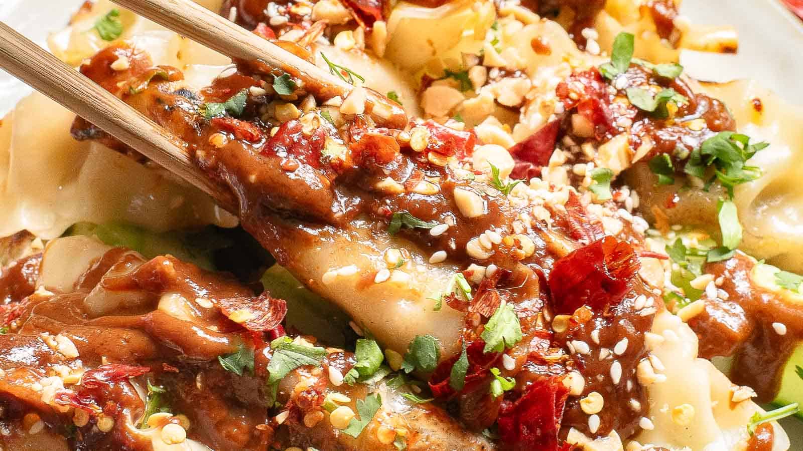 Close-up of dumplings topped with chili oil, crushed peanuts, sesame seeds, chopped herbs, and sauce, held by chopsticks.