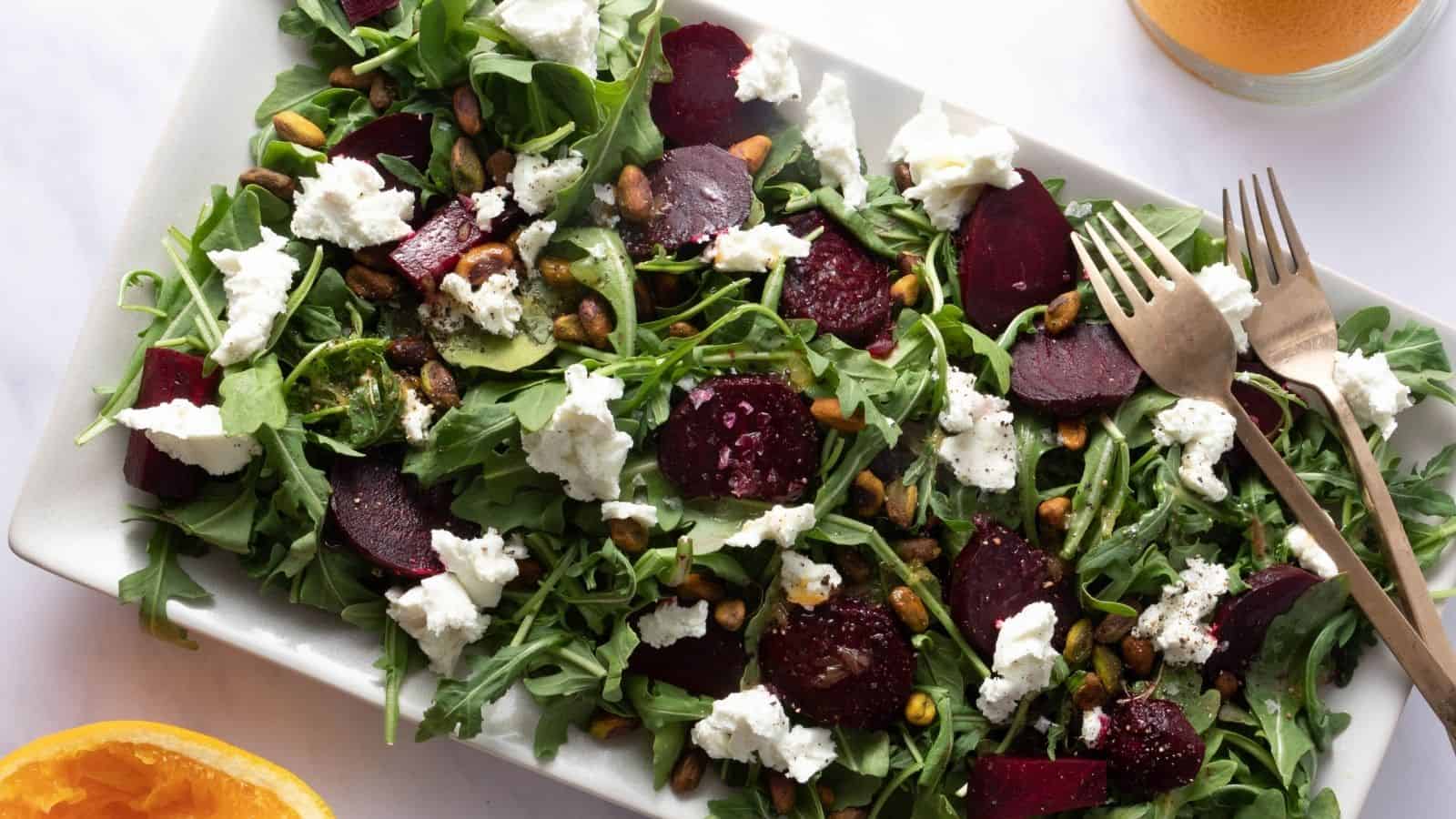 Rectangular plate with arugula, sliced beets, crumbled goat cheese, pistachios, and vinaigrette. Fork and knife on the side; partial glass of orange liquid is visible.