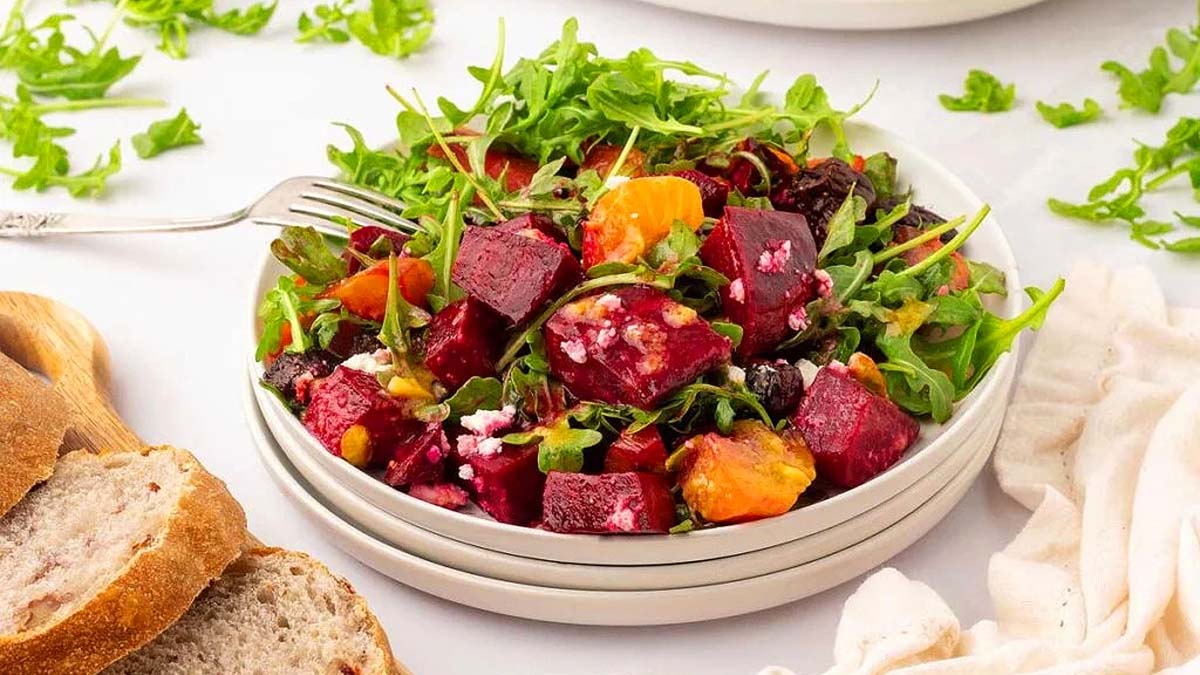 A plate of beet salad with arugula, orange slices, crumbled cheese, and nuts, served with slices of bread on the side.