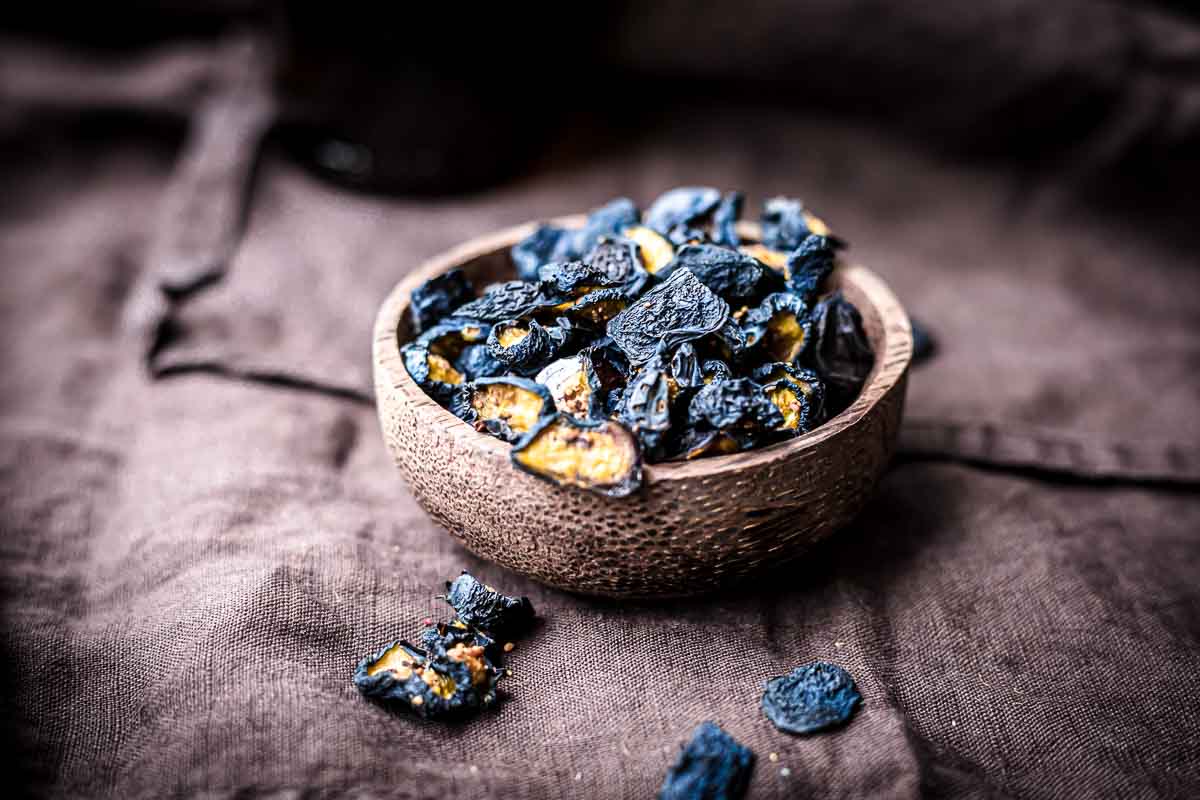 A wooden bowl filled with dried, dark purple or black fruit pieces sits on a brown fabric surface, with a few pieces scattered nearby.