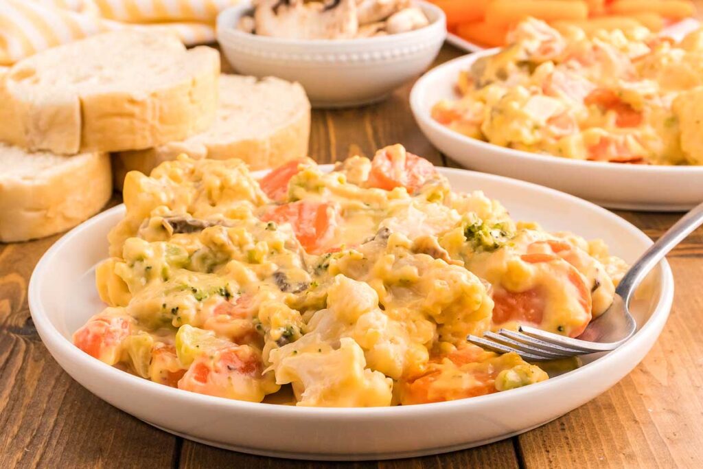 A plate of creamy chicken casserole with vegetables sits on a wooden table, accompanied by slices of bread and a bowl of mushrooms in the background.