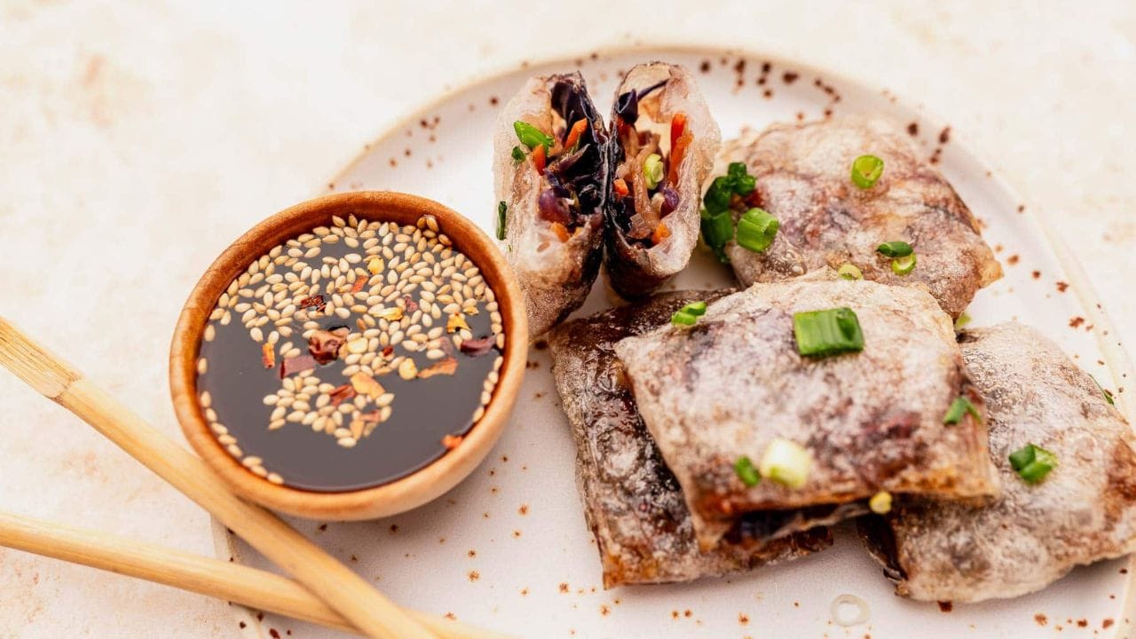 Plate with sliced crispy spring rolls filled with vegetables, accompanied by a small bowl of soy dipping sauce topped with sesame seeds and chili flakes, and a pair of chopsticks.