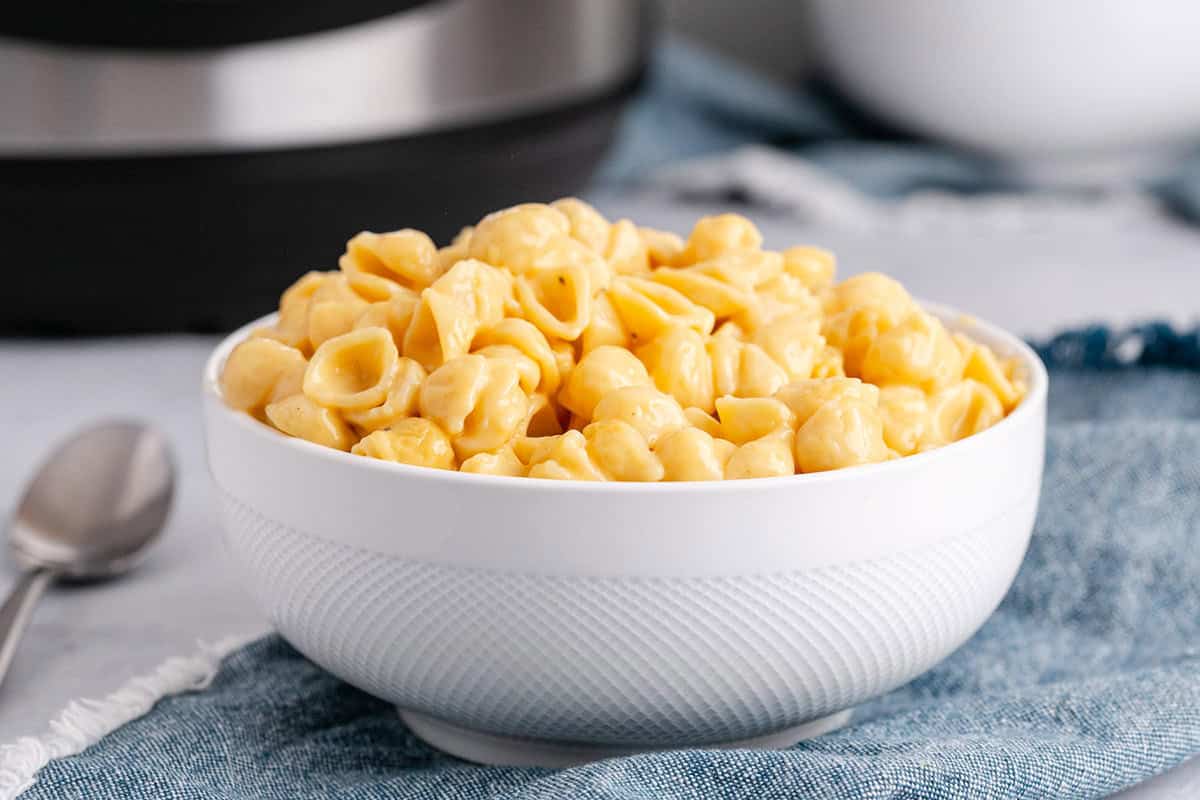 A white bowl filled with creamy macaroni and cheese pasta sits on a blue cloth next to a spoon.