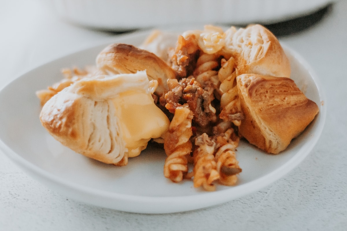 A white plate with spiral pasta in meat sauce surrounded by three pieces of flaky pastry, one showing melted cheese inside.