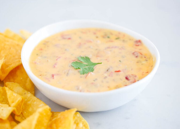 A white bowl of queso dip garnished with a cilantro leaf, next to a pile of tortilla chips on a white surface.