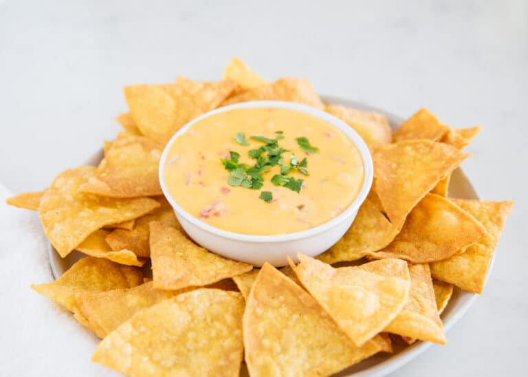 A bowl of cheese dip garnished with chopped herbs is surrounded by crispy tortilla chips on a plate.