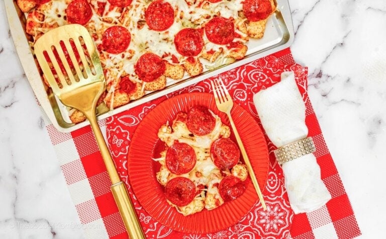 A plate with pepperoni pizza sits on a red checkered tablecloth next to a baking sheet of pizza, a gold spatula, and a white napkin wrapped with a jeweled ring.