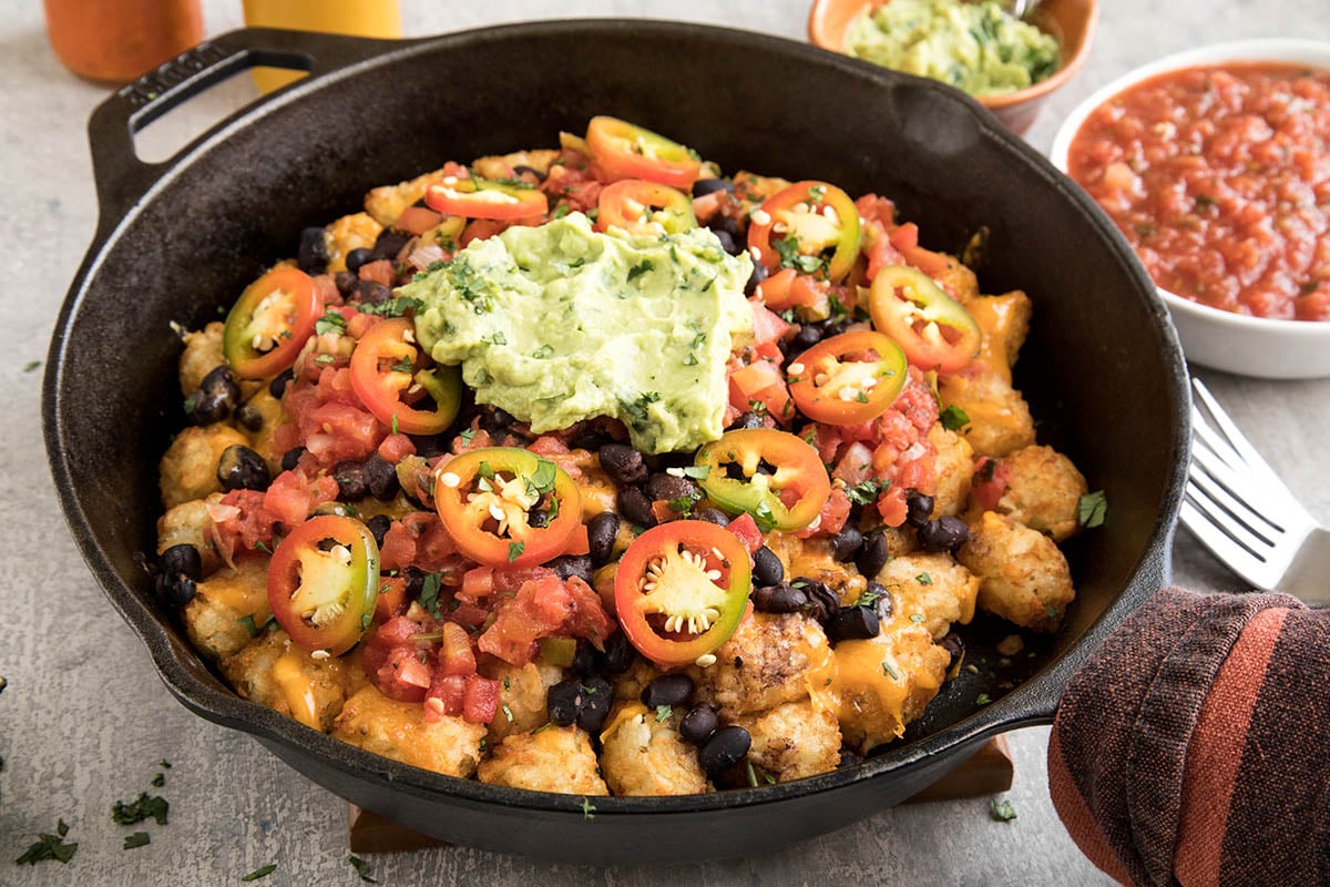 A cast iron skillet filled with tater tots topped with melted cheese, black beans, diced tomatoes, sliced jalapeรฑos, and a dollop of guacamole. Salsa and guacamole are in small bowls nearby.