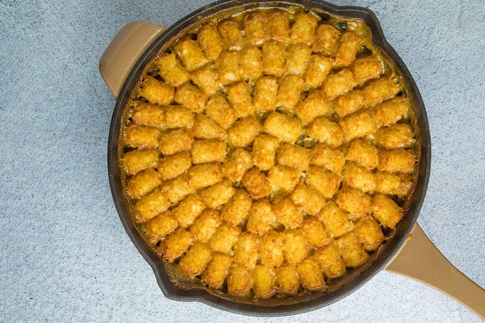 A skillet filled with tater tots arranged in a circular pattern over a casserole on a light blue surface.