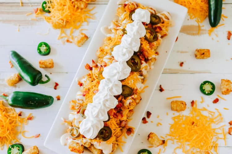Rectangular white plate with loaded tater tots topped with shredded cheese, jalapeรฑos, bacon, and sour cream, surrounded by jalapeรฑos, tater tots, and cheese on a white surface.