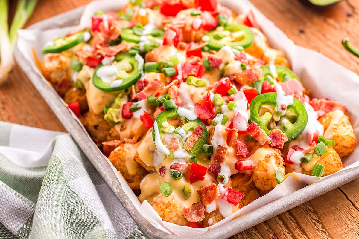 A tray of loaded tater tots topped with melted cheese, sliced jalapeรฑos, bacon, diced tomatoes, green onions, and a drizzle of white sauce.