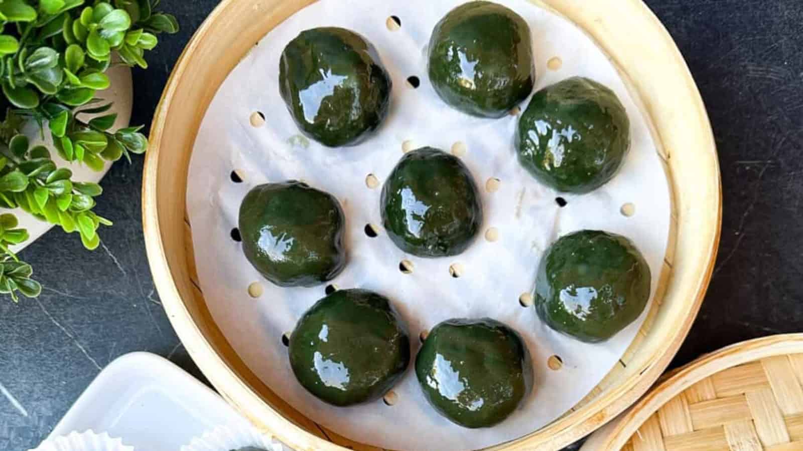 Eight round green dumplings are arranged on parchment paper in a bamboo steamer basket, with a leafy plant visible nearby.