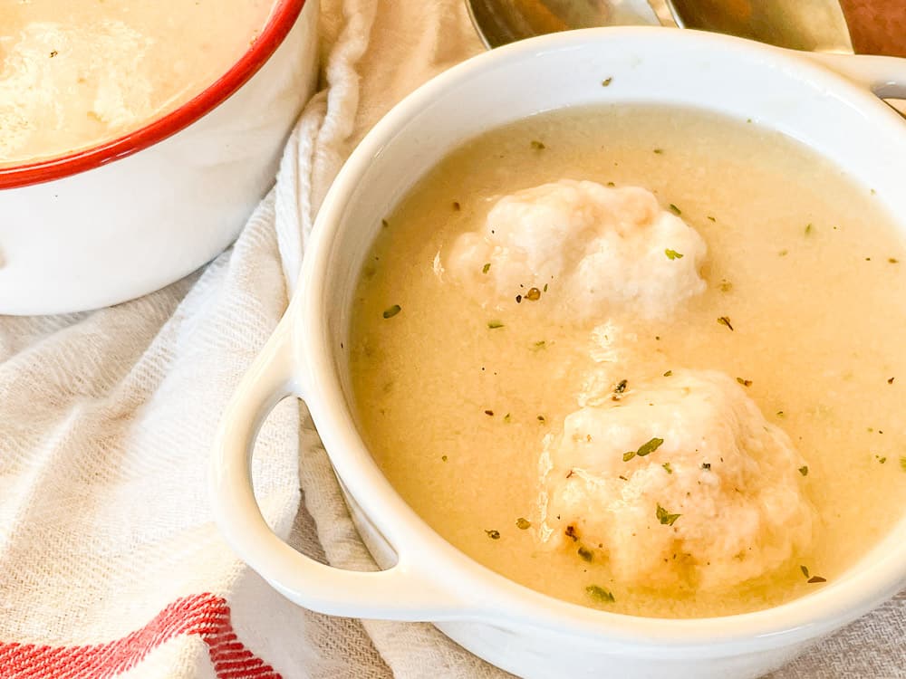 A white bowl filled with creamy soup and two dumplings, garnished with herbs, sits on a striped towel next to a pot.