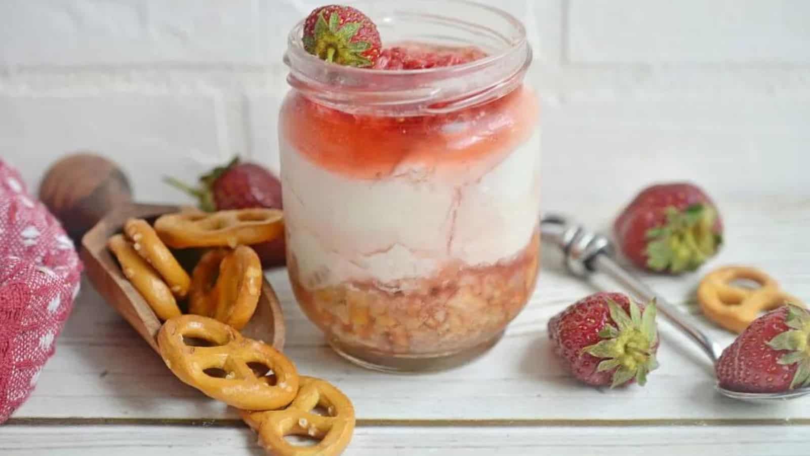 A jar filled with layered cheesecake, topped with strawberry sauce and a whole strawberry, surrounded by pretzels and fresh strawberries on a white wooden surface.