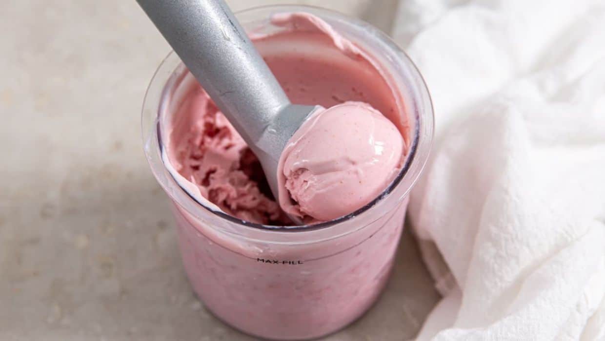 A metal ice cream scoop rests in a container of pink ice cream, with a scoop of ice cream on the utensil. A white cloth is beside the container.