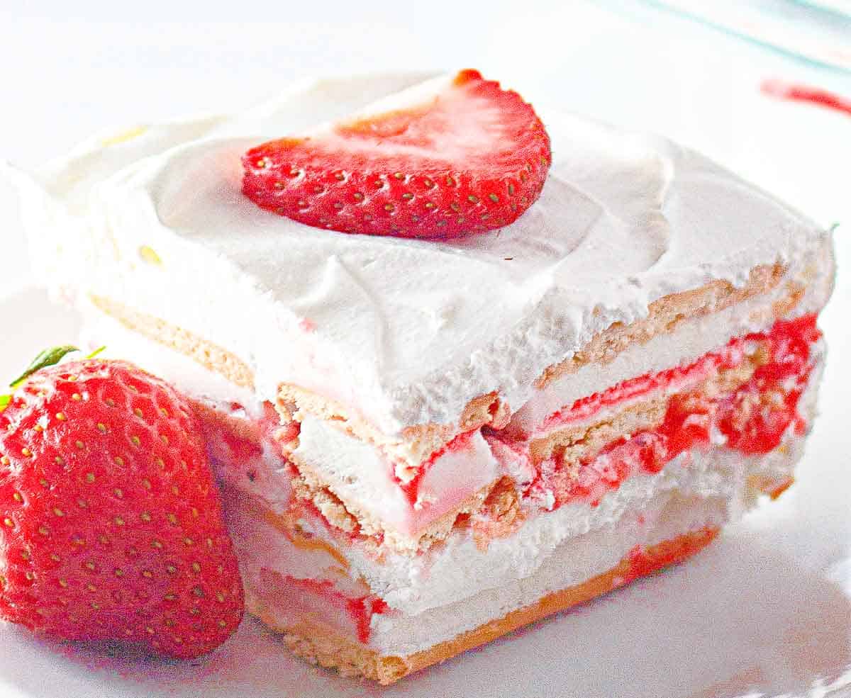 A slice of strawberry icebox cake with layers of whipped cream and cookies, topped with a strawberry slice, on a white plate next to a whole strawberry.