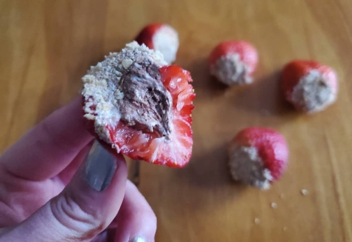 A hand holds a half-eaten strawberry filled with chocolate and coated in crushed crumbs, with more similarly prepared strawberries on a wooden surface in the background.