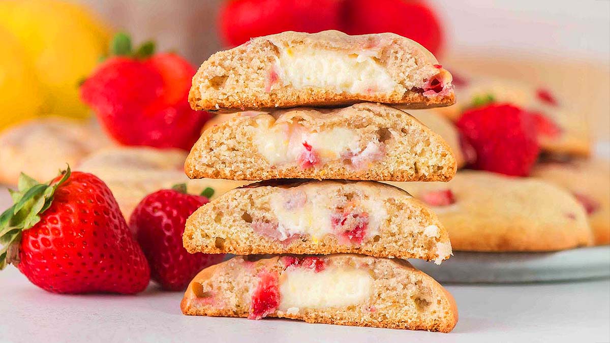 Stack of cookies with creamy centers and pieces of strawberries, with whole strawberries and blurred fruit in the background.